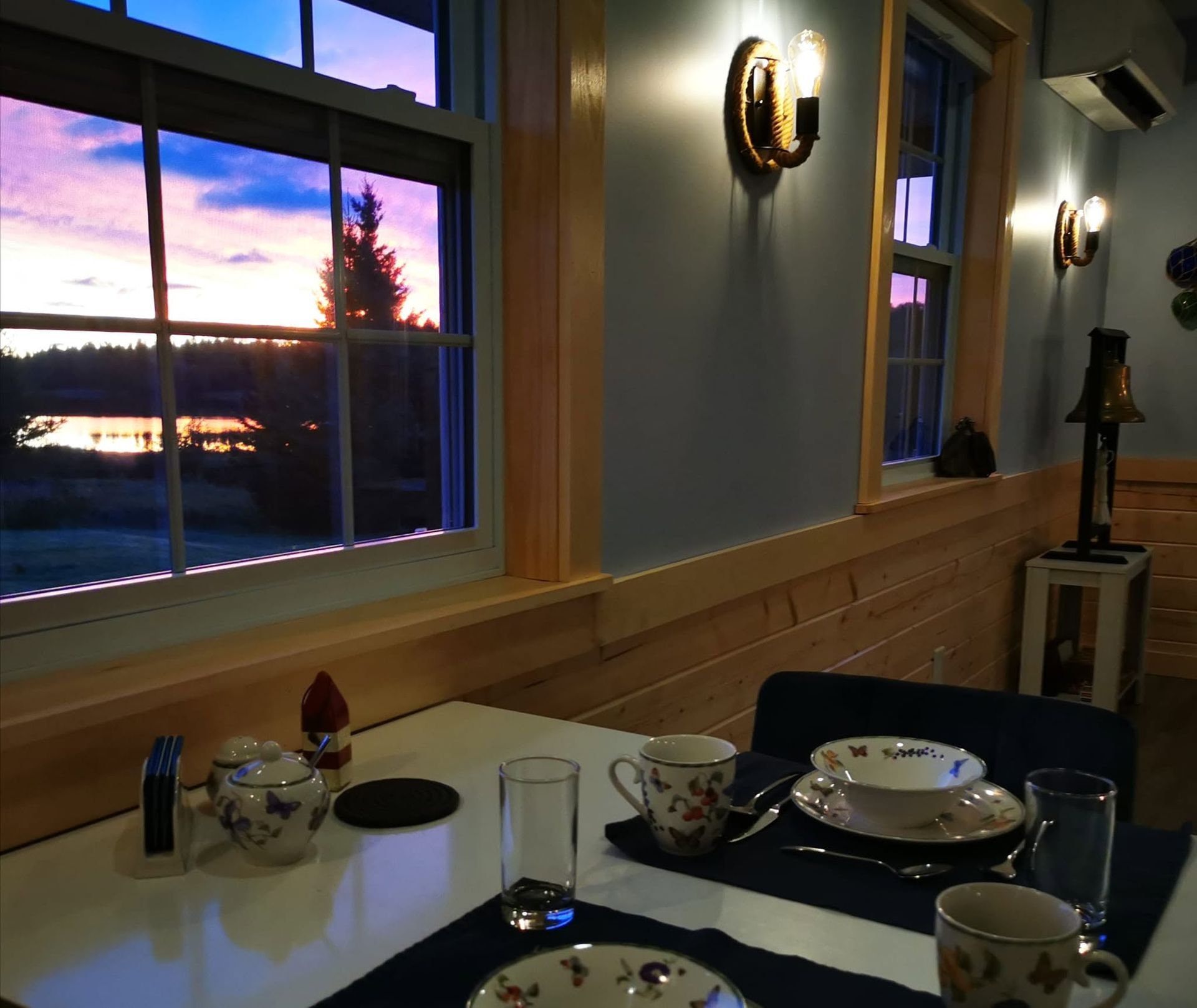 Gezellige restauranttafel bij grote ramen met uitzicht op de zonsondergang, met blauw servies en warme wandlampen.