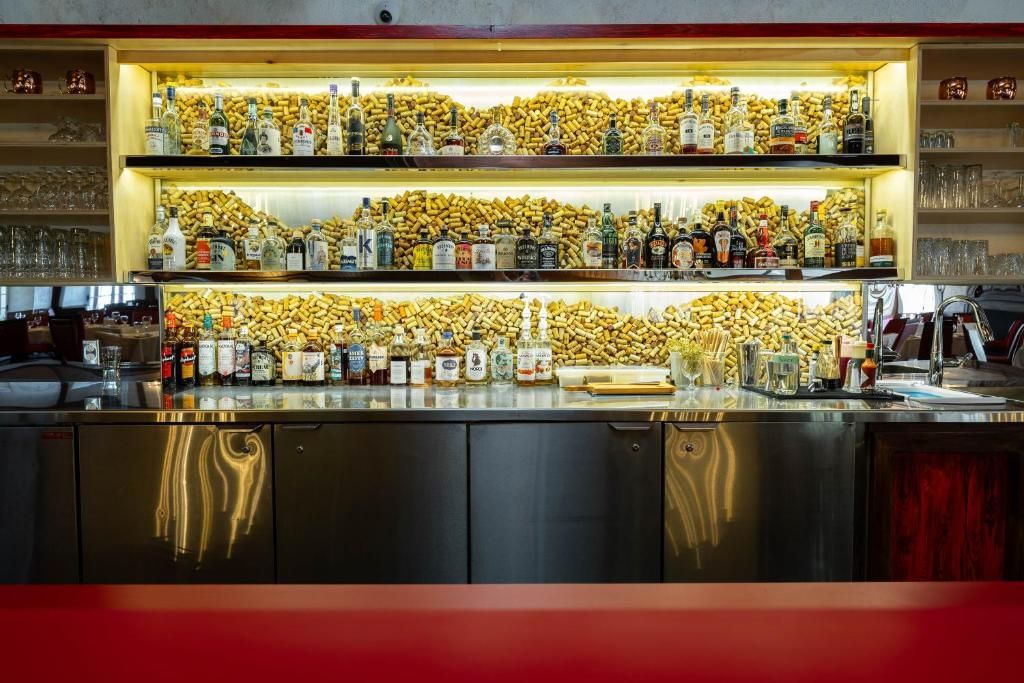Een bar met achtergrondverlichting, rijen flessen en glazen, een donkere toonbank en rode zitplaatsen ervoor.