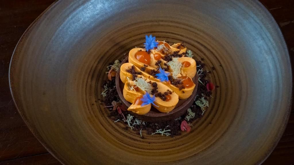 Een gastronomisch dessert met sinaasappelschijfjes en blauwe eetbare bloemen op een donker bord met een brede rand.