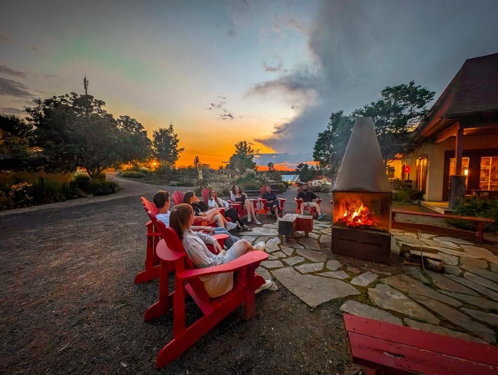 Mensen ontspannen rond een vuurplaats bij Le Baluchon Éco-villégiature bij zonsondergang op een stenen terras met rode stoelen en een nabijgelegen blokhut.