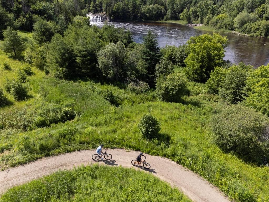 Twee fietsers rijden over een kronkelig grindpad langs een rivier, omgeven door weelderige groene bomen en struiken.