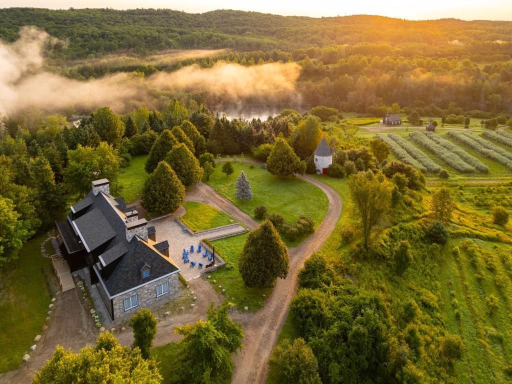 Luchtfoto van een landelijk huis en een kronkelende zandweg in weelderige groene velden bij zonsopgang, met mist over de heuvels.