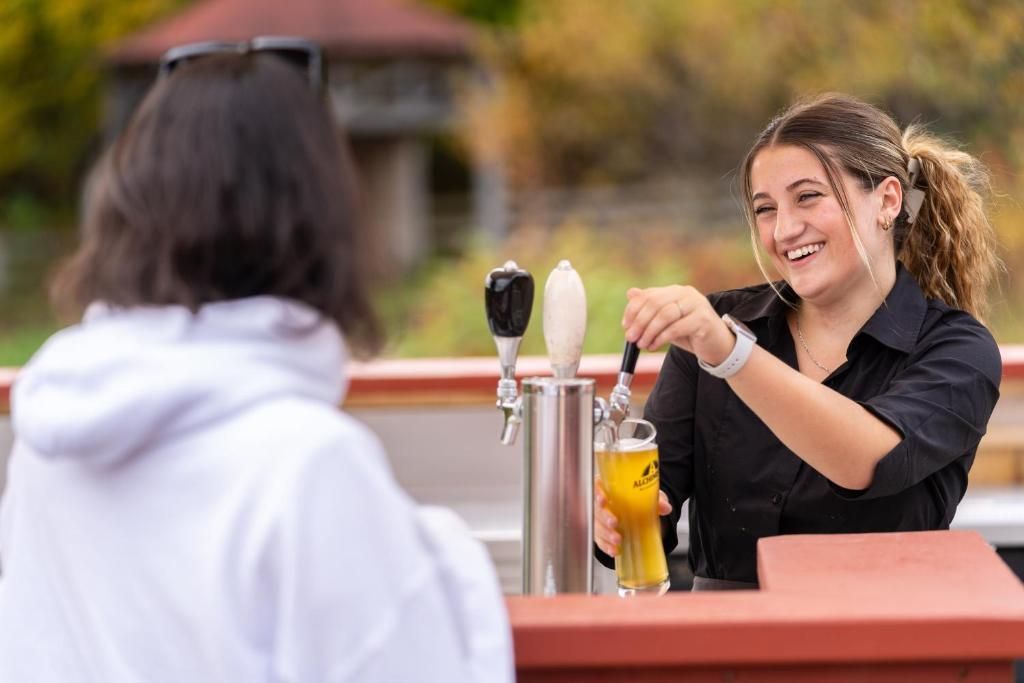 Twee mensen zitten te praten aan een tafeltje buiten, een van hen serveert een drankje naast metalen dispensers.