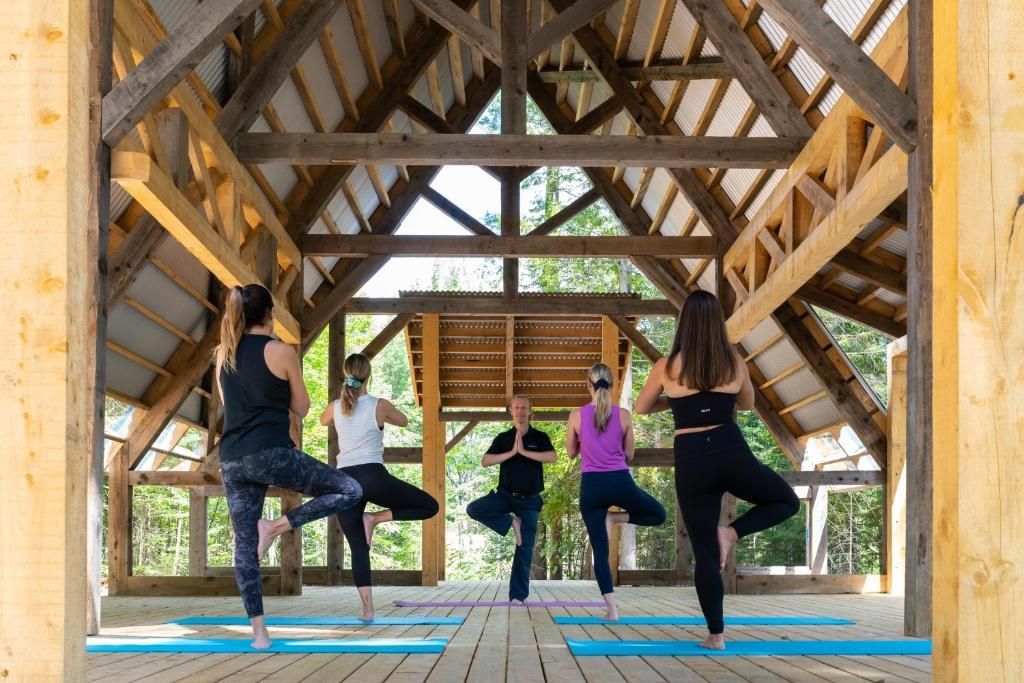 Vier mensen doen de boomhouding op yogamatten in een houten paviljoen buiten.