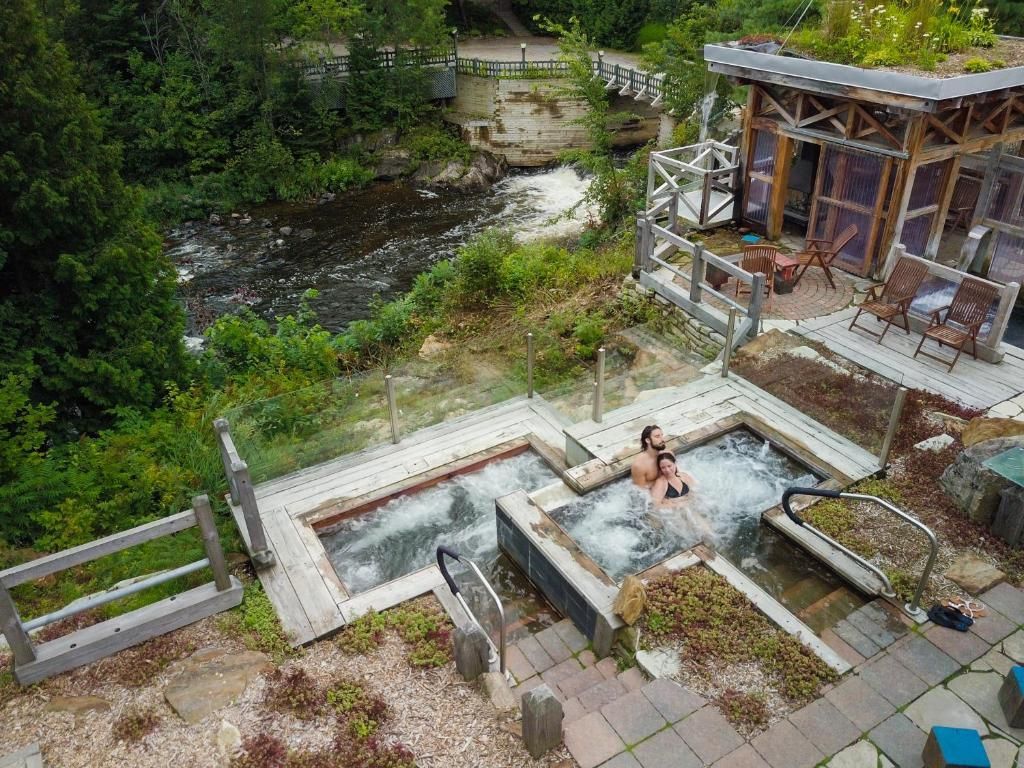 Een stenen jacuzzi naast een beekje in het bos, met twee mensen die ontspannen op een terras bij een rustiek gebouw.