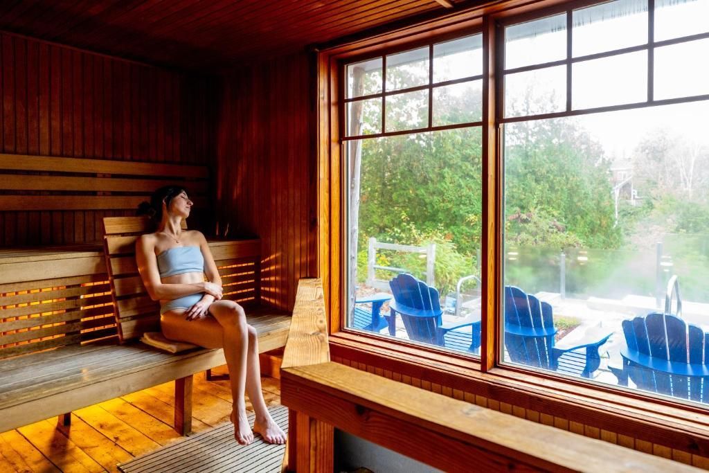 Vrouw zit in een houten saunalounge naast een groot raam met uitzicht op blauwe stoelen en groen.