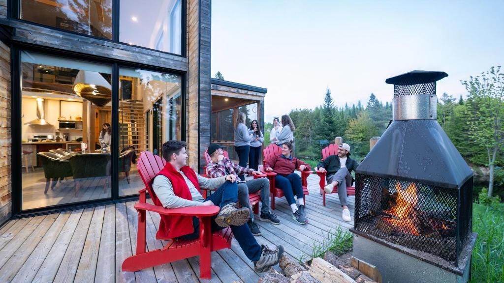 Mensen ontspannen op rode stoelen rond een vuurplaats naast een modern huis in de schemering.