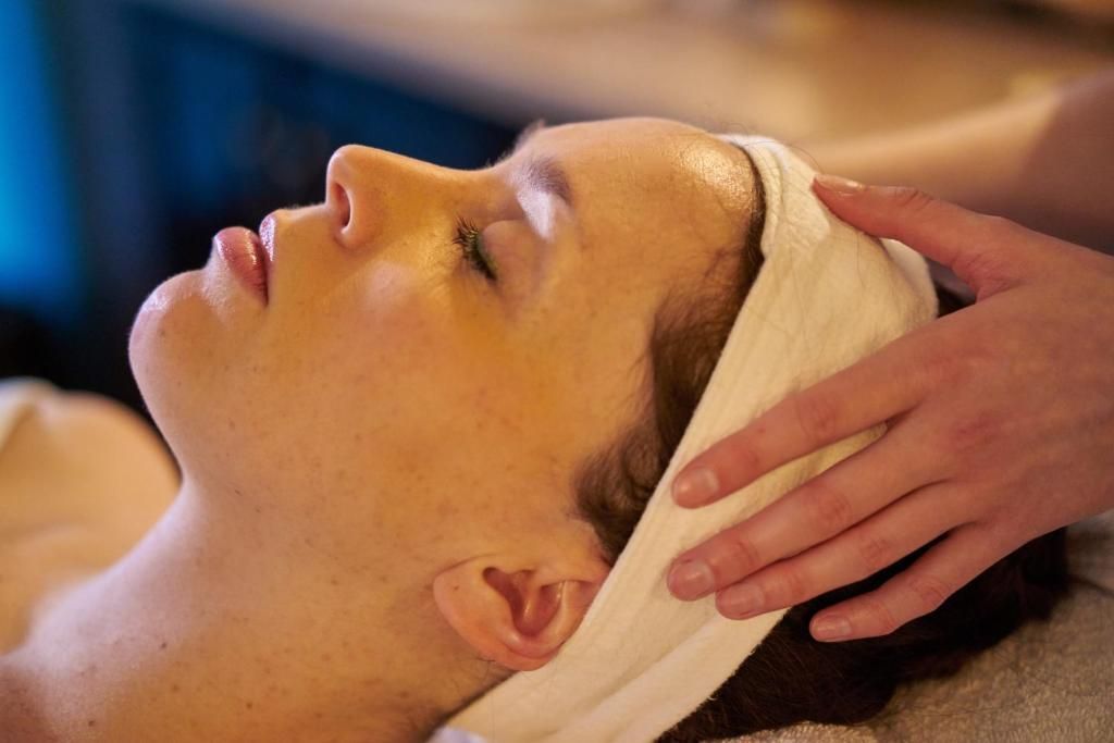 Een persoon krijgt een gezichtsmassage met gesloten ogen, draagt een witte hoofdband en een handdoek.