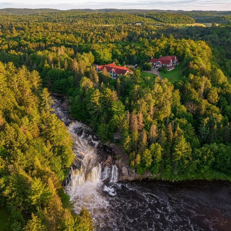 Luchtfoto van een waterval naast een dicht bos en gebouwen met rode daken aan een rivier.