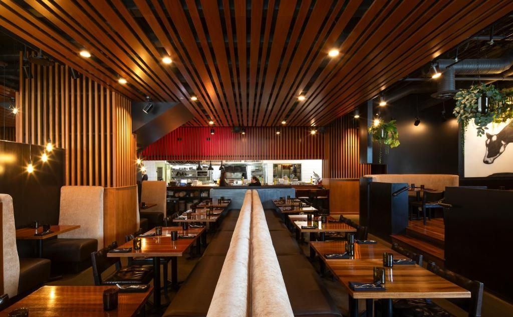 Modern restaurantinterieur met houten plafond, warme verlichting en netjes gedekte tafels langs een centrale gang.