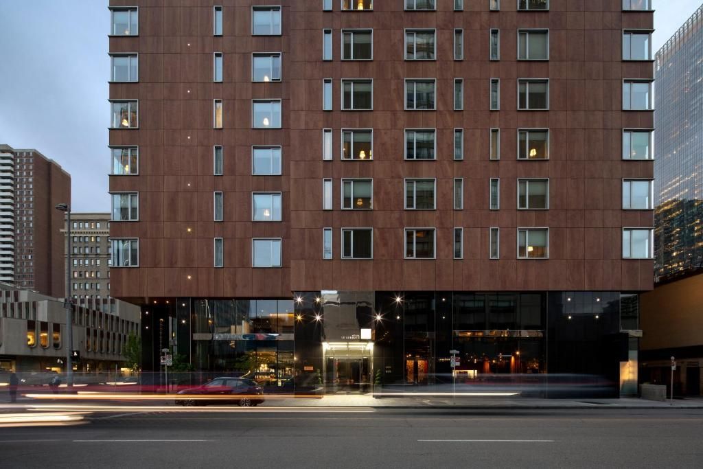 Een hoog, bruin bakstenen appartementencomplex met verlichte winkels op de begane grond in de schemering, gezien vanaf de overkant van de straat.