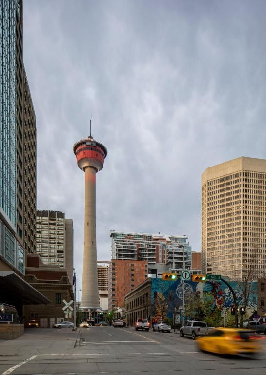 Stadsstraat met de Calgary Tower, hoge gebouwen en een gele taxi die onder een bewolkte hemel voorbijrijdt.