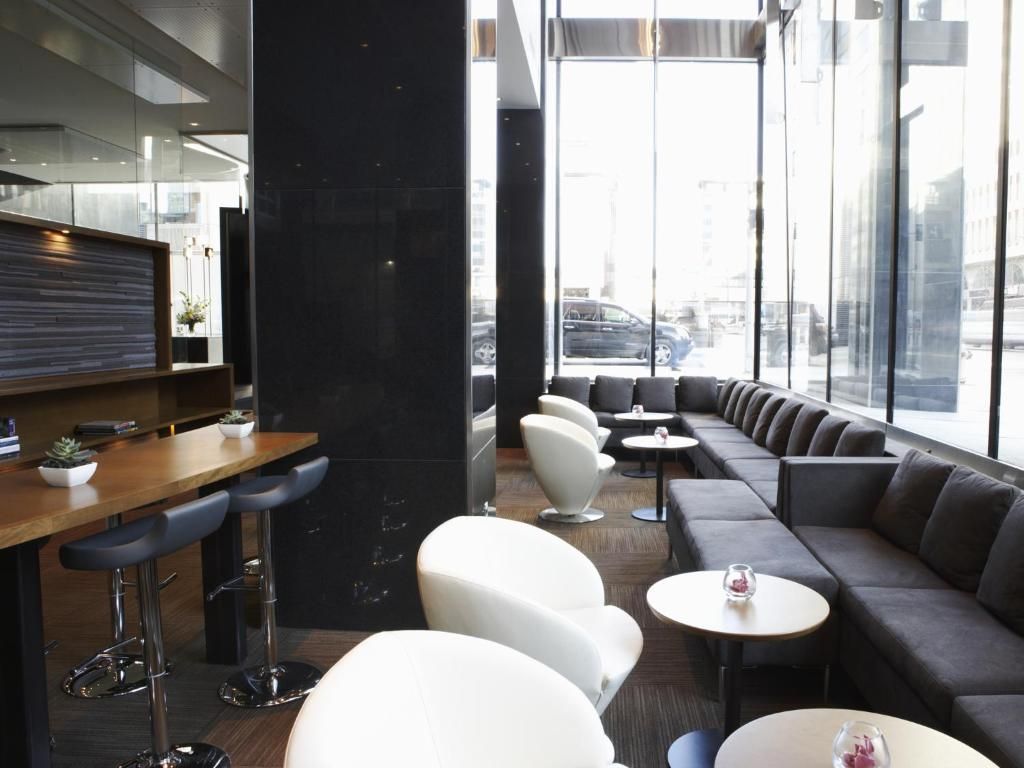 Moderne woonkamer met donkere banken, ronde witte tafels, barkrukken en grote ramen die veel licht binnenlaten.