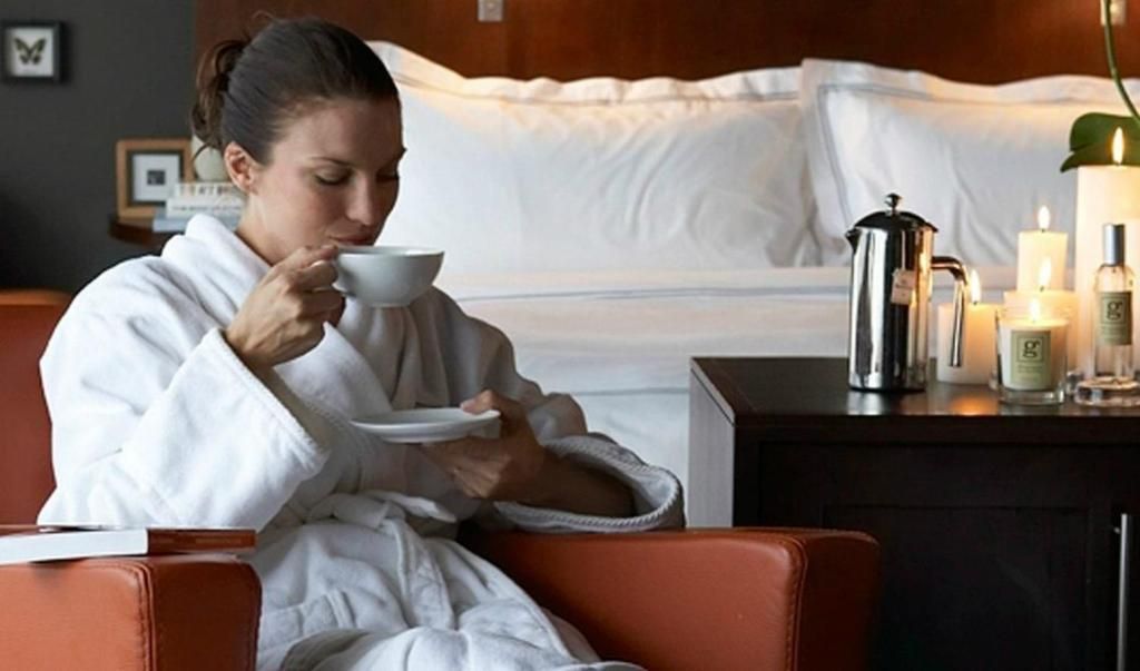 Vrouw in een witte ochtendjas drinkt uit een kopje in een hotelkamer, zittend naast een bed met kaarsen en een koffiepot.