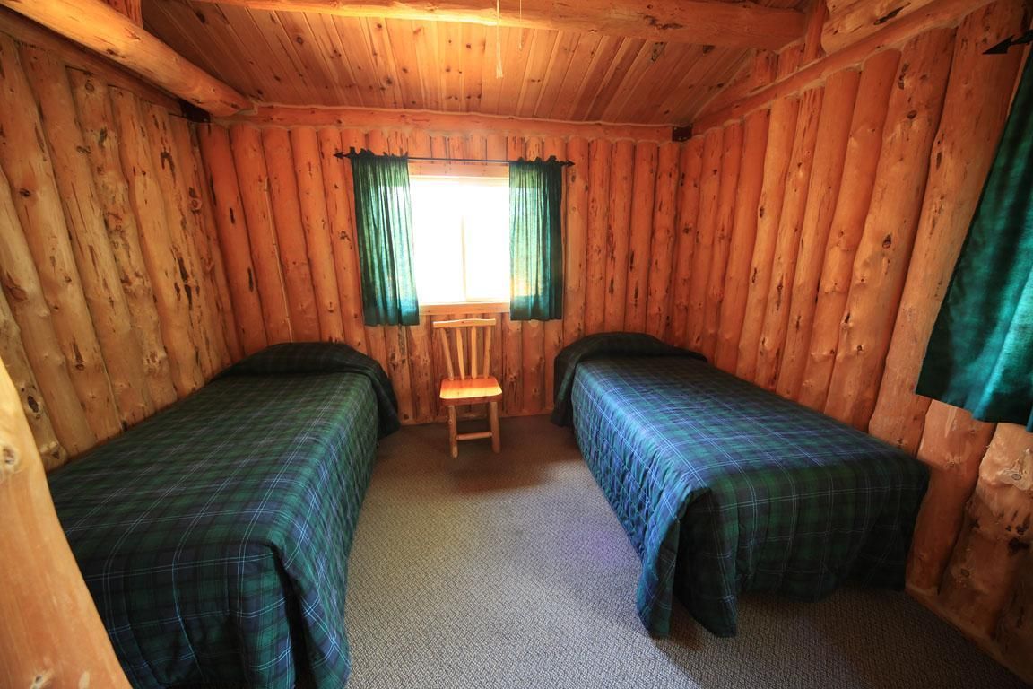 Rustieke slaapkamer in een blokhut met twee eenpersoonsbedden, een houten stoel en een raam met groene gordijnen.