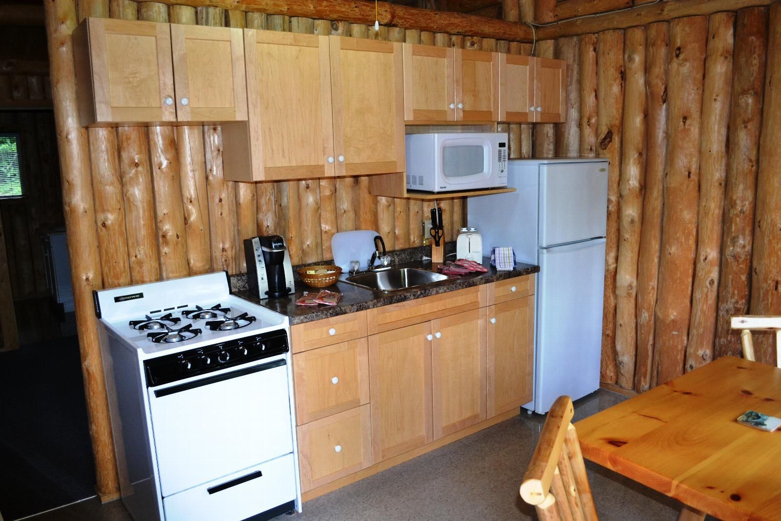 Kleine, rustieke keuken met houten wanden, wit fornuis, magnetron, koelkast en houten kastjes en tafel.