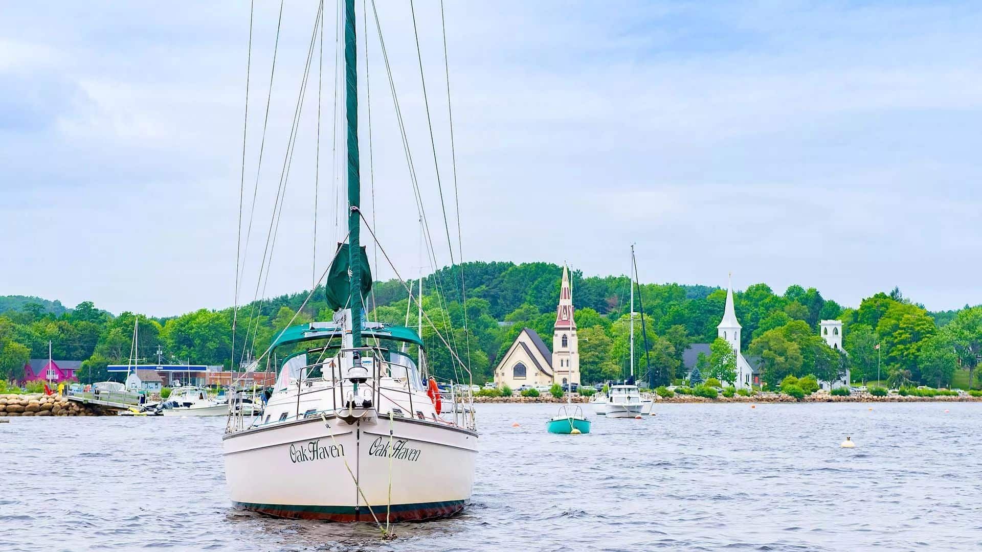 Zeilboot op woelig water nabij een groene kustlijn en een witte kerk onder een bewolkte hemel.