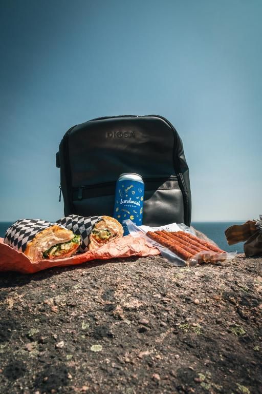 Een picknick met snacks en een blauw blikje op een rots, met een zwarte rugzak tegen een blauwe lucht.