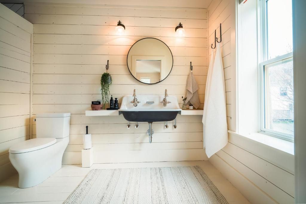 Lichte badkamer met witte houten lambrisering, zwart wastafelmeubel, ronde spiegel en vrijstaand bad bij het raam.