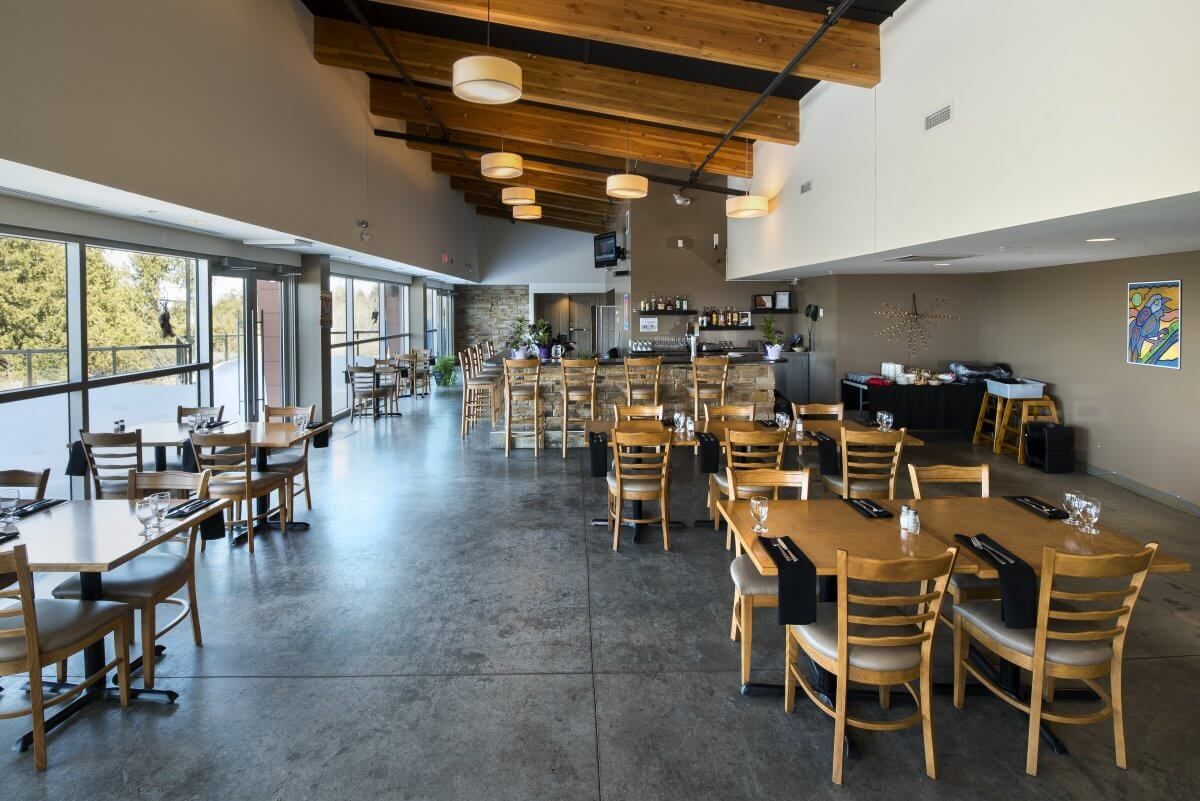 Ruime, moderne restaurantzaal met houten tafels, stoelen en grote ramen aan één kant.