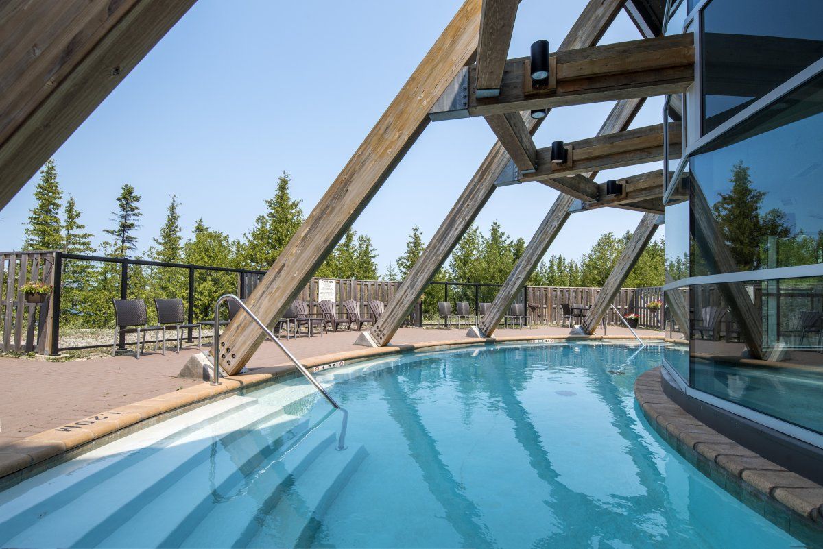 Modern resortzwembad met blauw water, houten balken en een glazen gebouw dat ernaast wordt weerspiegeld.