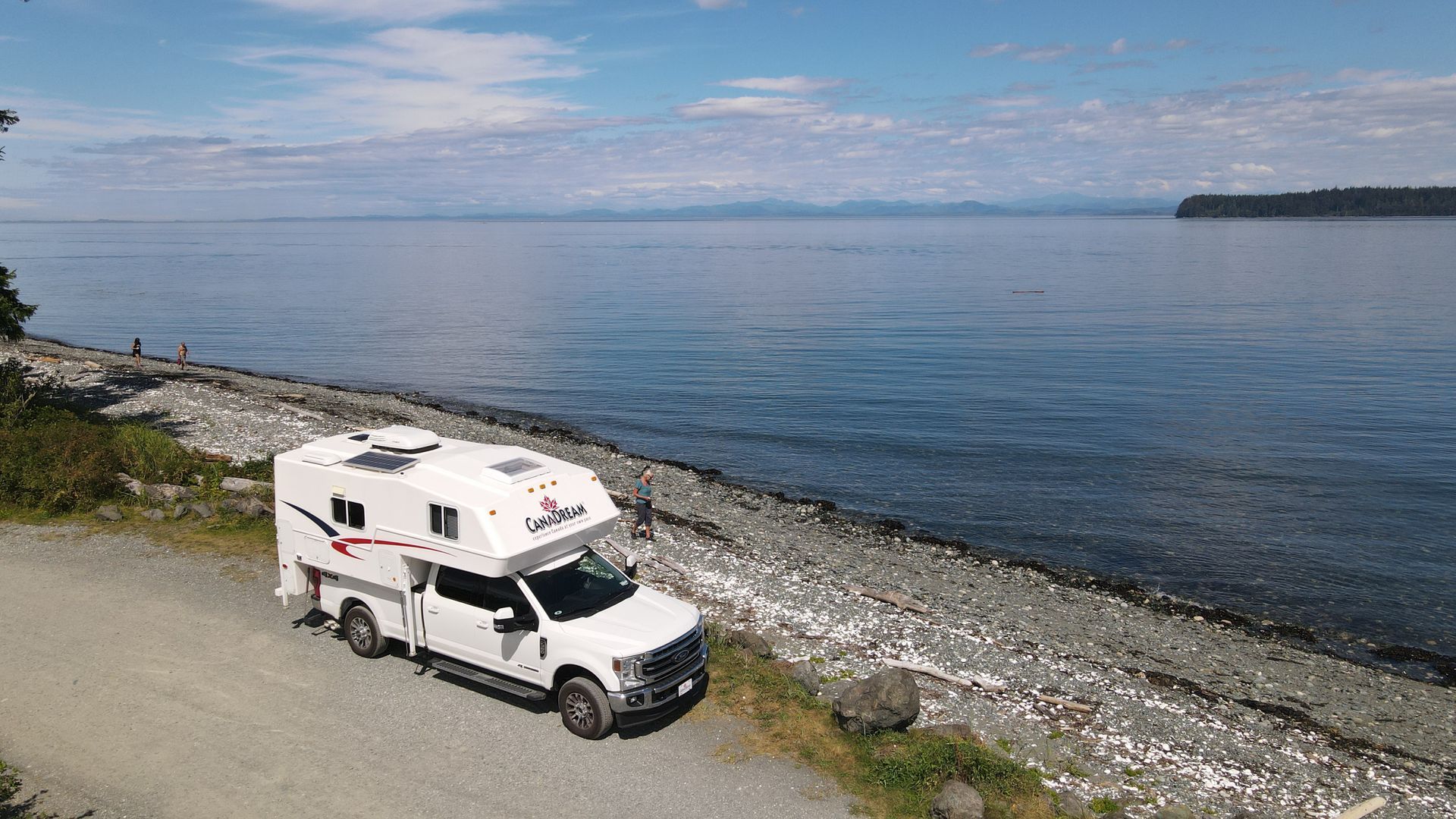 Witte Maxi Travel 4x4 Camper geparkeerd op een kiezelstrand naast rustig blauw water