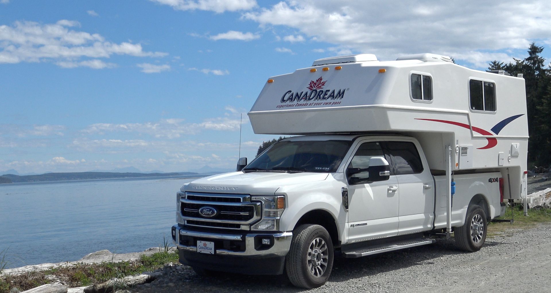 Witte Maxi Travel 4x4 Camper geparkeerd op een kiezelstrand naast kalm water