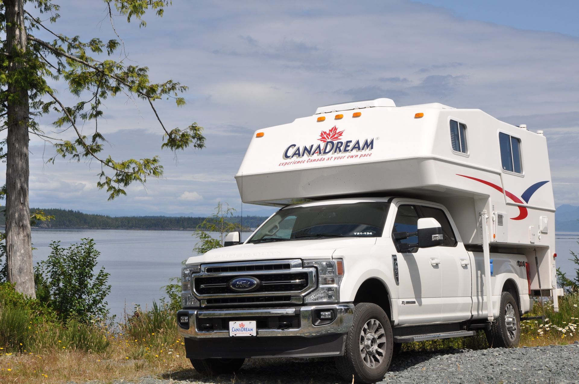 Witte Maxi Travel 4x4 Camper geparkeerd aan een oever onder een bewolkte hemel, met bomen en water