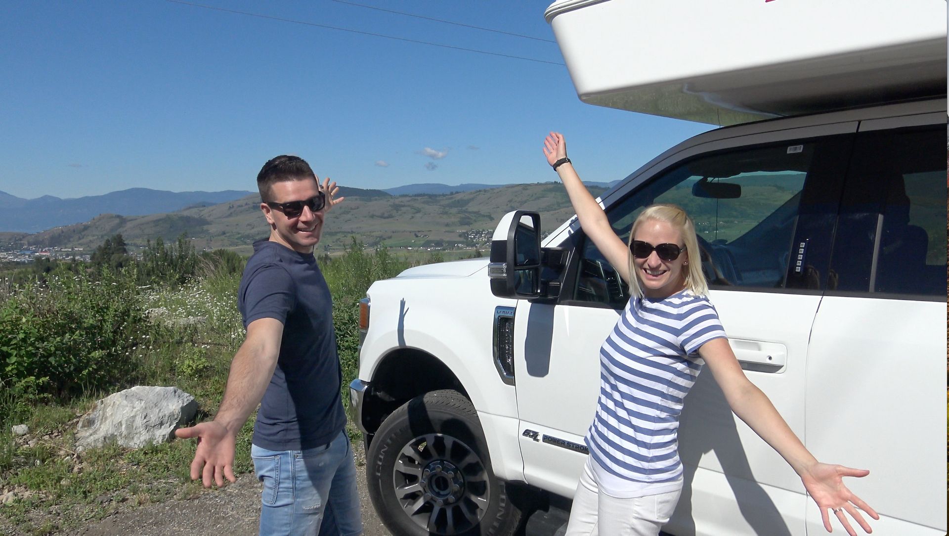 Twee mensen poseren naast een witte CanaDream Maxi Travel Camper in een zonnig berglandschap