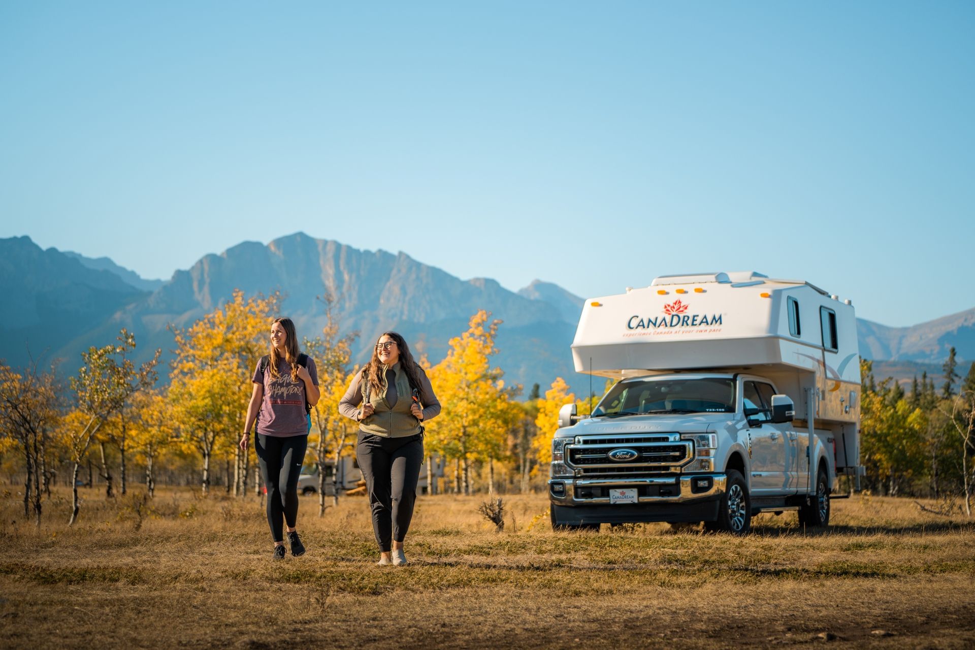 Twee vrouwen lopen naast een witte Maxi Travel 4x4 Camper langs herfstbomen