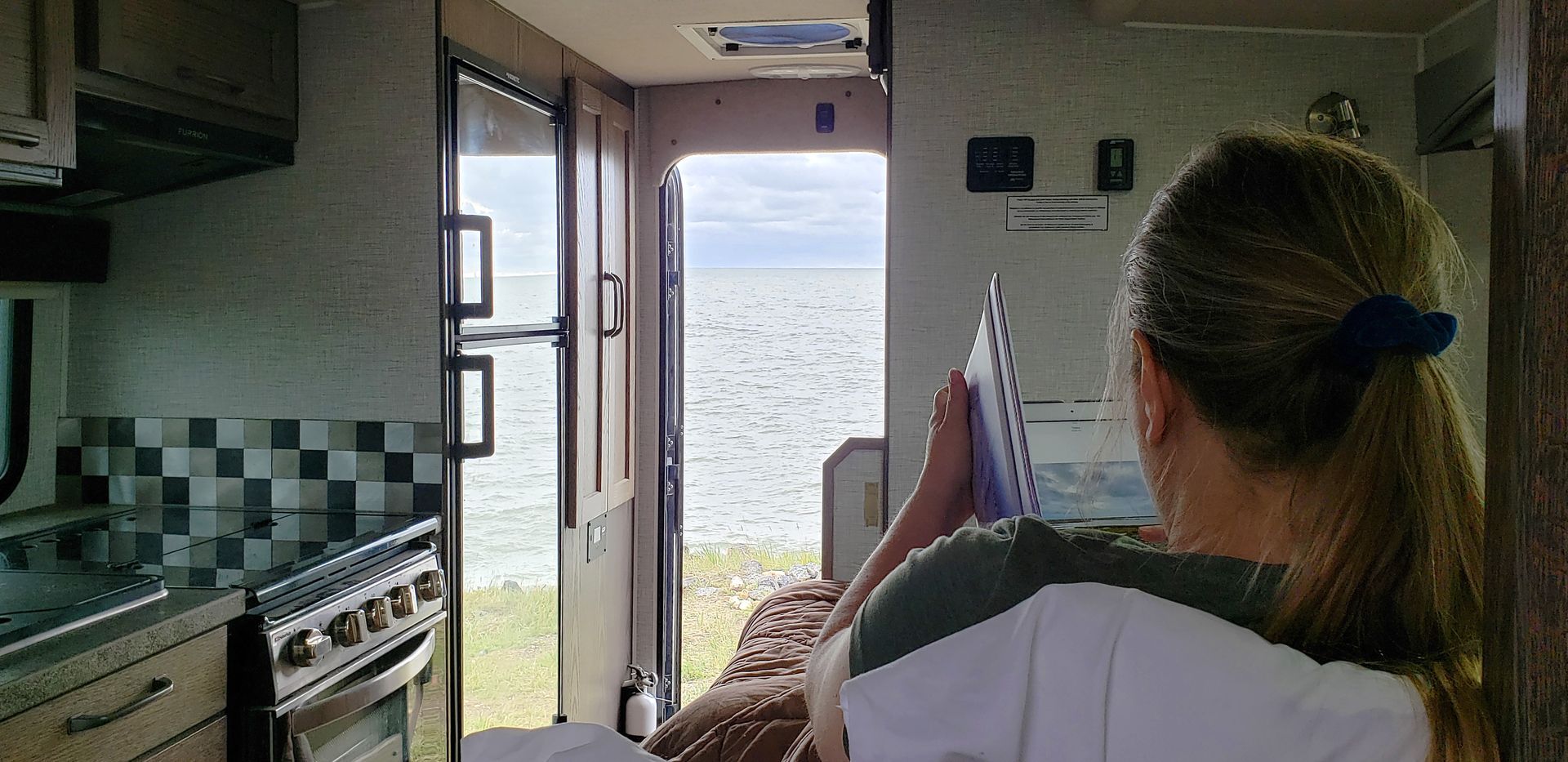 Persoon zit in een Maxi Travel 4x4 Camper en kijkt door de open deur naar een helder uitzicht op zee.