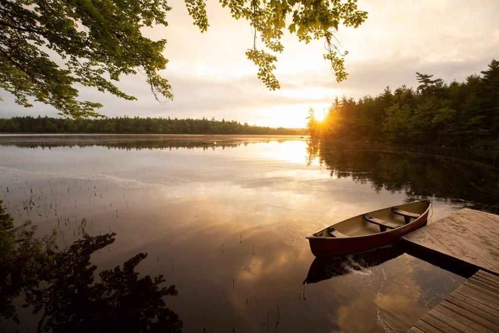 Zonsondergang boven een kalm meer met een kano aangemeerd aan een steiger en bomen die in het water weerspiegeld worden.