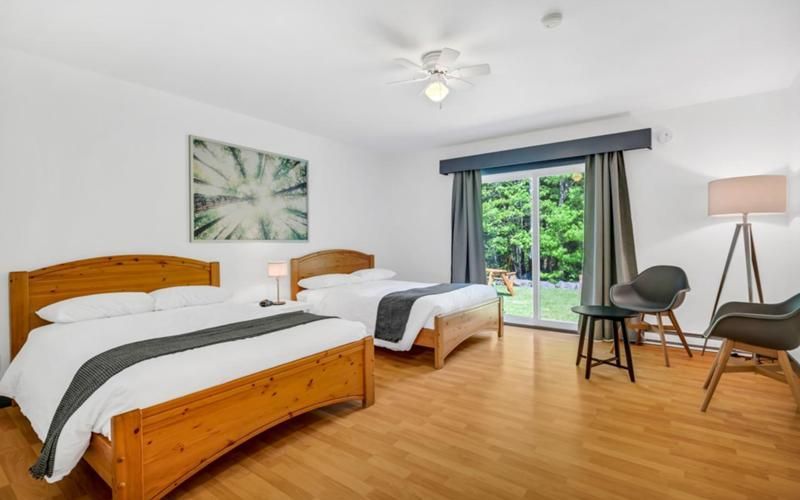 Lichte slaapkamer met twee houten bedden, grijze gordijnen, een glazen schuifdeur en een rieten stoel naast een staande lamp.