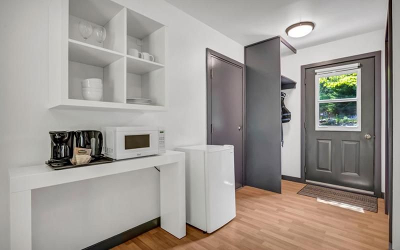 Lichte entreehal met witte inbouwplanken, een klein bureau, een minikoelkast en een grijze voordeur op een houten vloer.