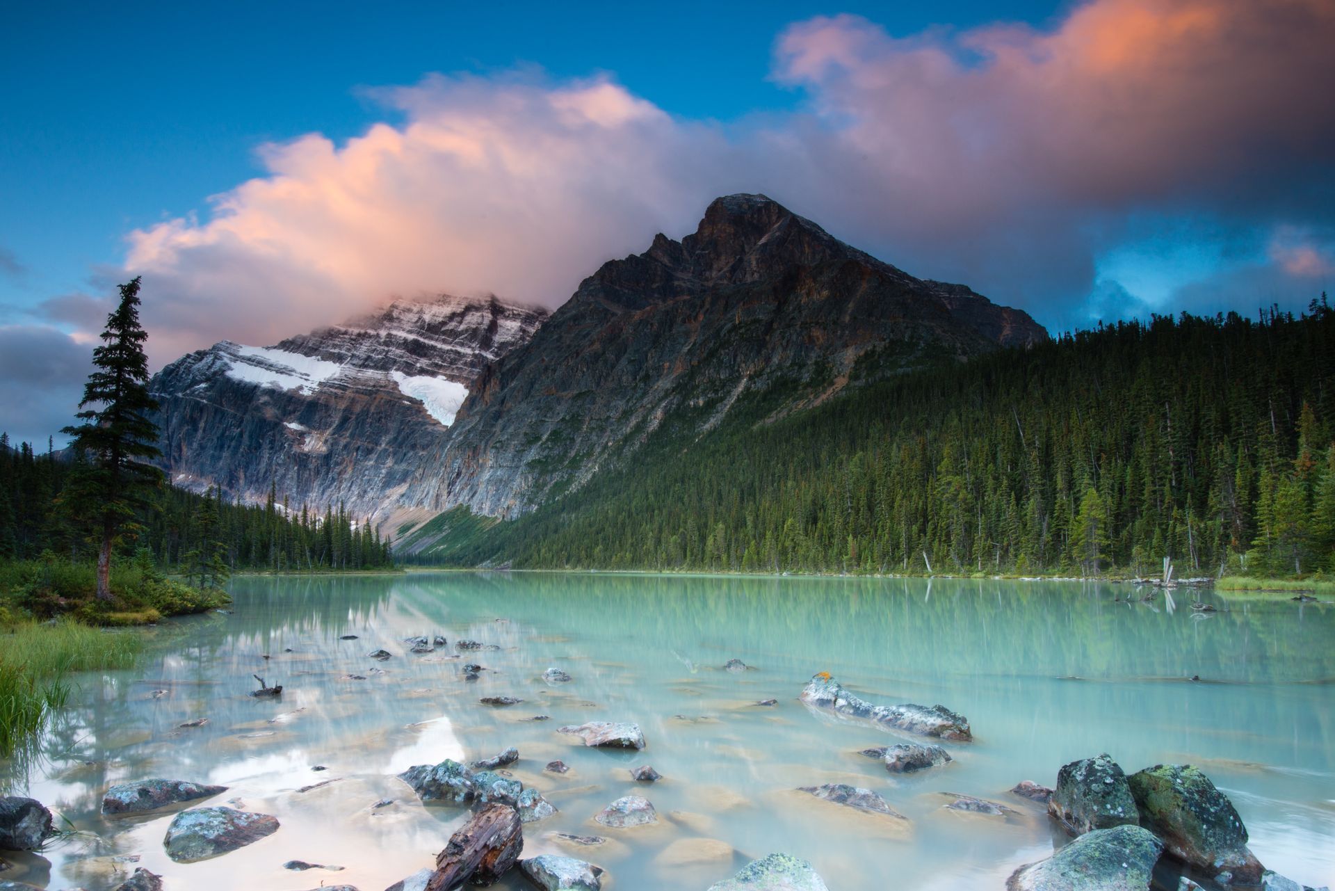 Bergmeer voor Mount Edith Cavell in Jasper National Park met turquoise water, dennenbomen, rotsachtige oever en roze wolken bij zonsondergang.