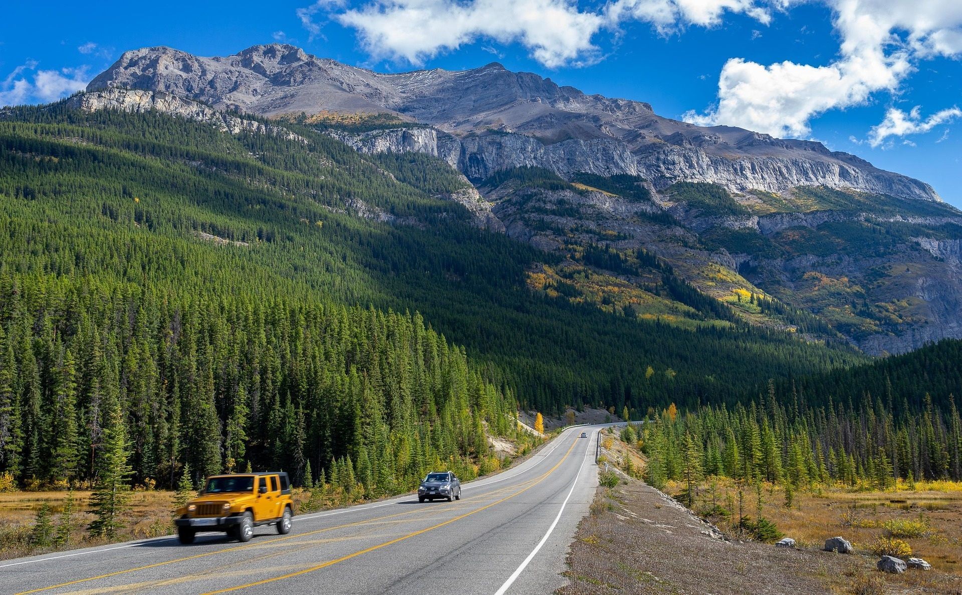 Bergweg langs Mount Robson, Canada met een gele Jeep, een auto, bos en rotsachtige bergtoppen onder een blauwe hemel