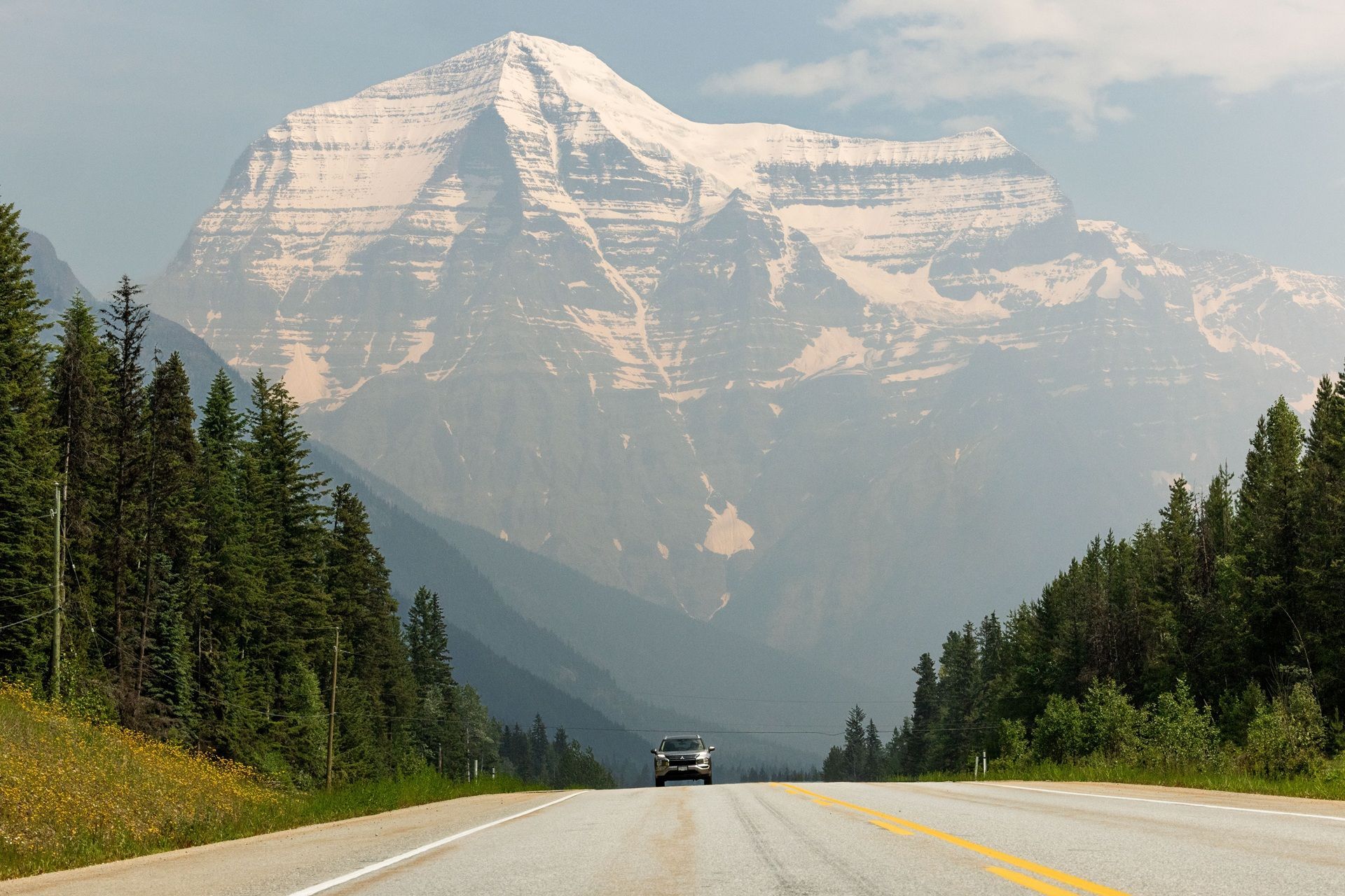 Een auto rijdt over een weg door dennenbossen richting een met sneeuw bedekte bergtop van Mount Robson onder een wazige hemel.