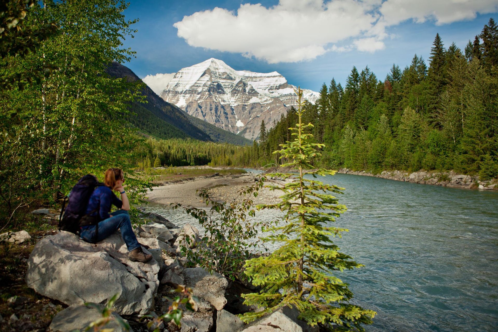 Wandelaars rusten uit bij een rivier bij de besneeuwde toppen van Mount Robson, dennenbomen en een gedeeltelijk bewolkte hemel.