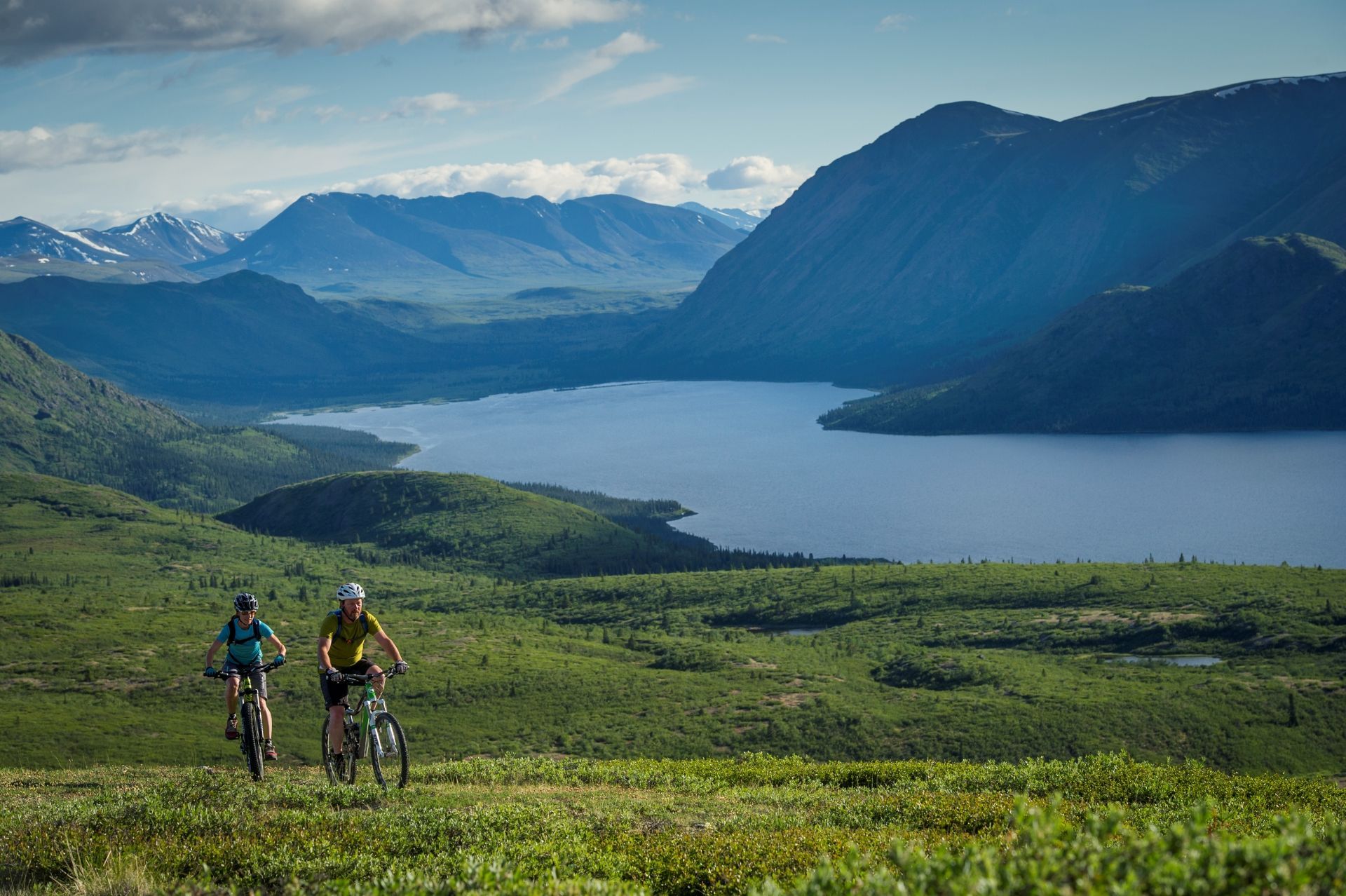 ©Government of Yukon Twee fietsers op een groen pad in Yukon, Canada met uitzicht op een meer en bergen