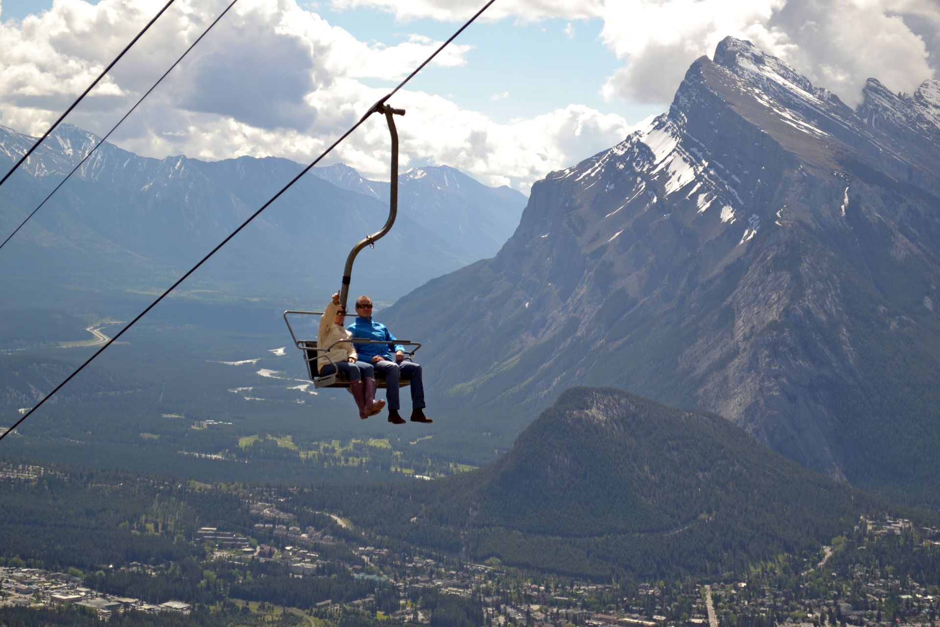 Twee mensen nemen een skilift naar Mount Norquay in Banff National Park boven een bergdal met besneeuwde toppen en wolken.