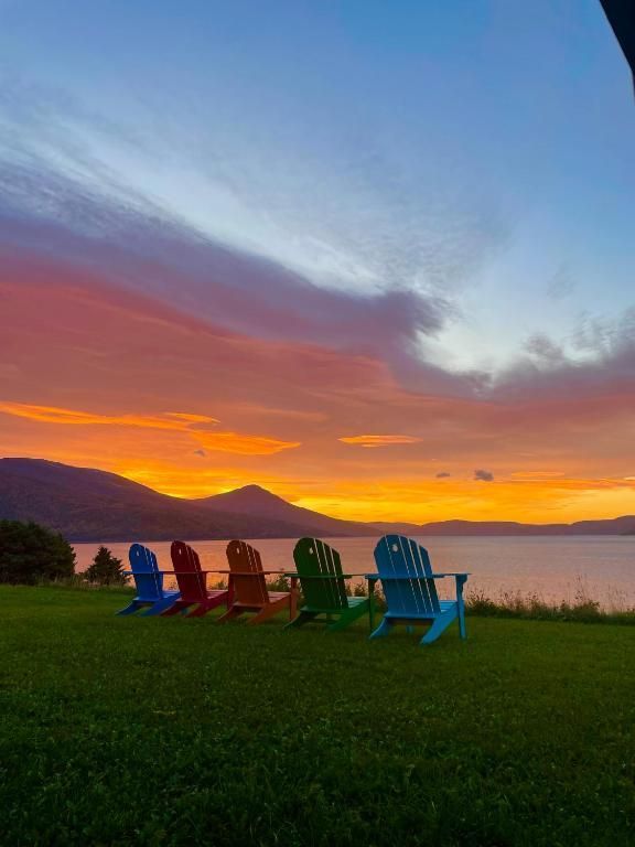 Een rij kleurrijke Adirondack-stoelen op het gras bij zonsondergang, met de bergen op de achtergrond en een gloeiende oranje hemel.