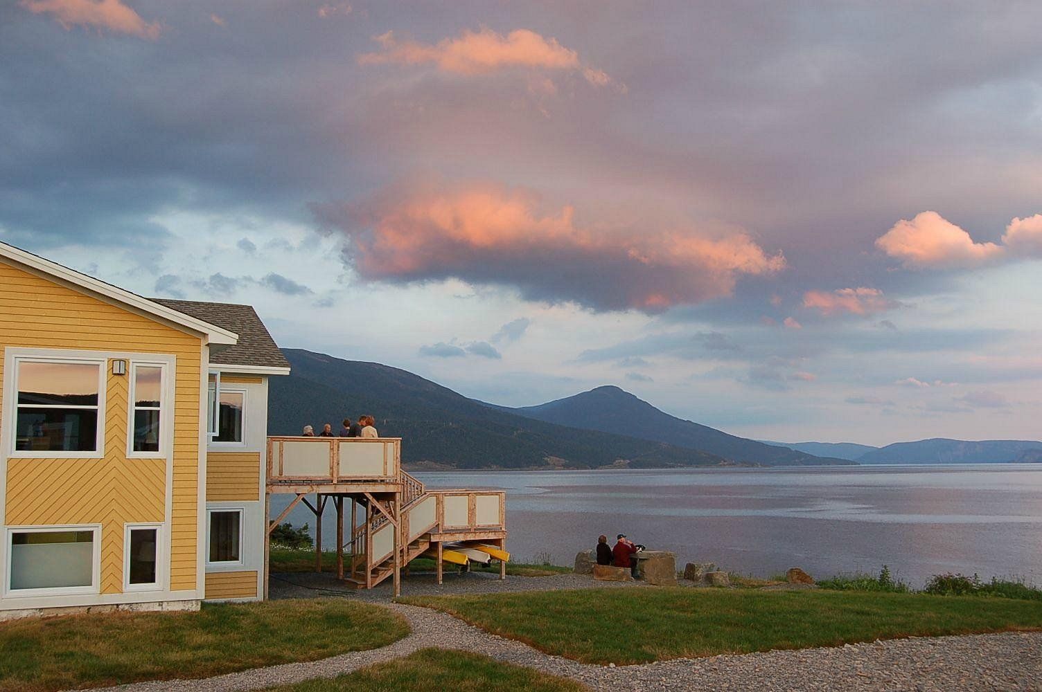 Geel huis met uitzicht op een rustige baai bij zonsondergang, met mensen die op het grasveld zitten.