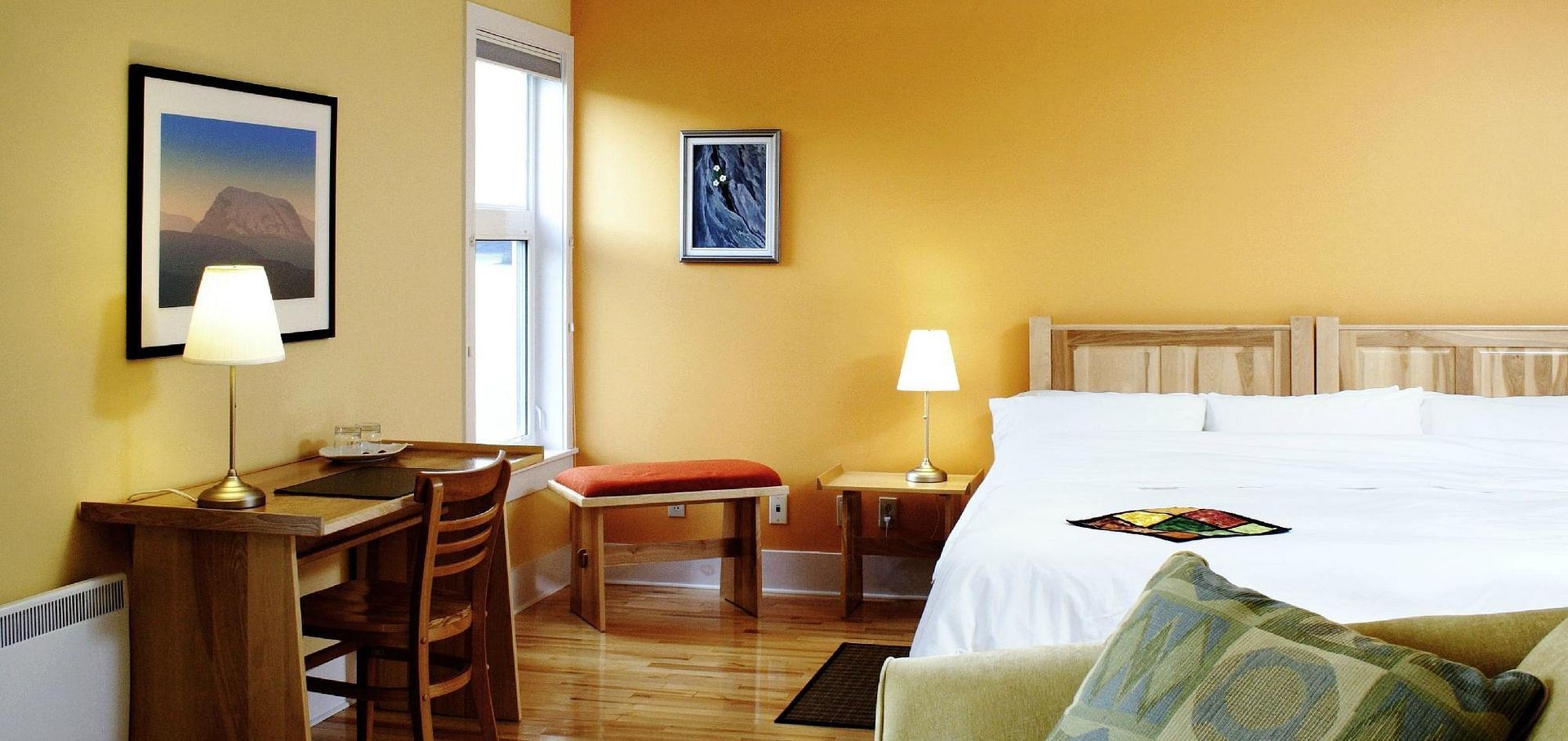 Gezellige hotelkamer met een bed, bureau, stoel, lamp en muurdecoratie in warme gele tinten.