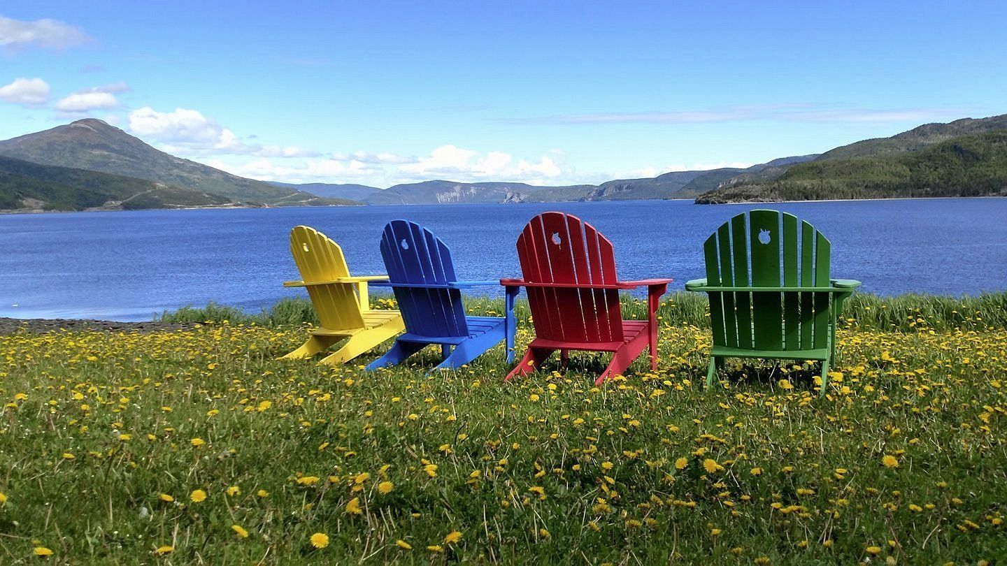 Kleurrijke Adirondack-stoelen op een grasrijke oever van een meer met bergen en blauw water op de achtergrond.