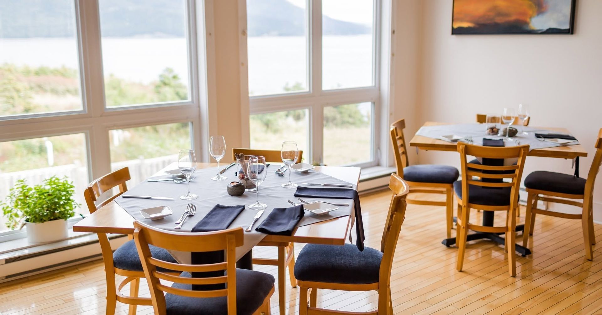 Zonnige eetkamer met houten tafels en stoelen naast grote ramen met uitzicht op het water.