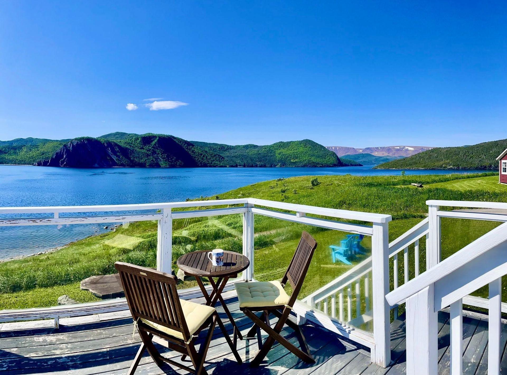 Terrasstoelen en een tafel bieden uitzicht op een blauwe baai, groene heuvels en een heldere hemel vanaf een terras aan zee.
