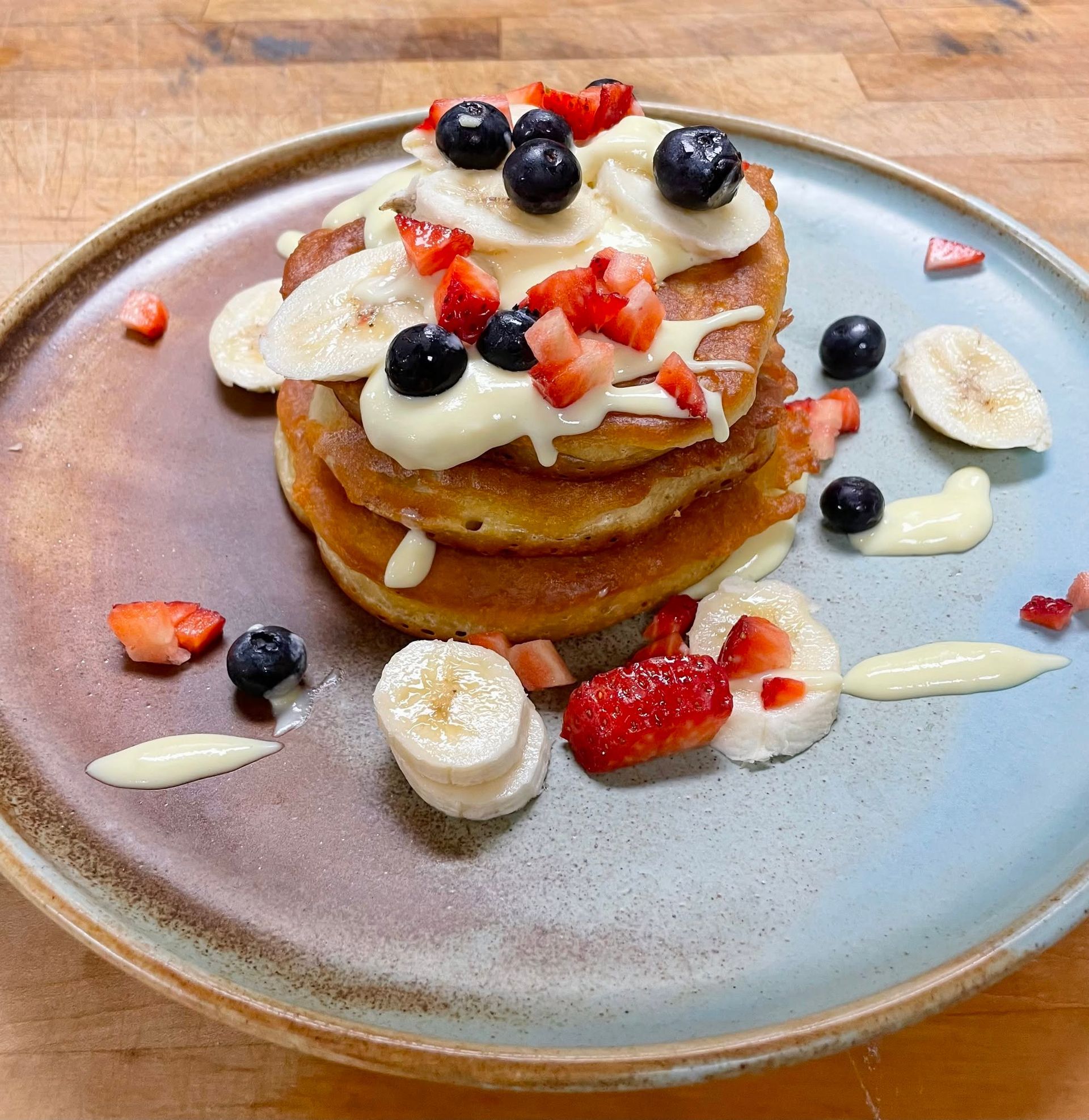 Een stapel pannenkoeken met slagroom, aardbeien, bosbessen en plakjes banaan op een bord.