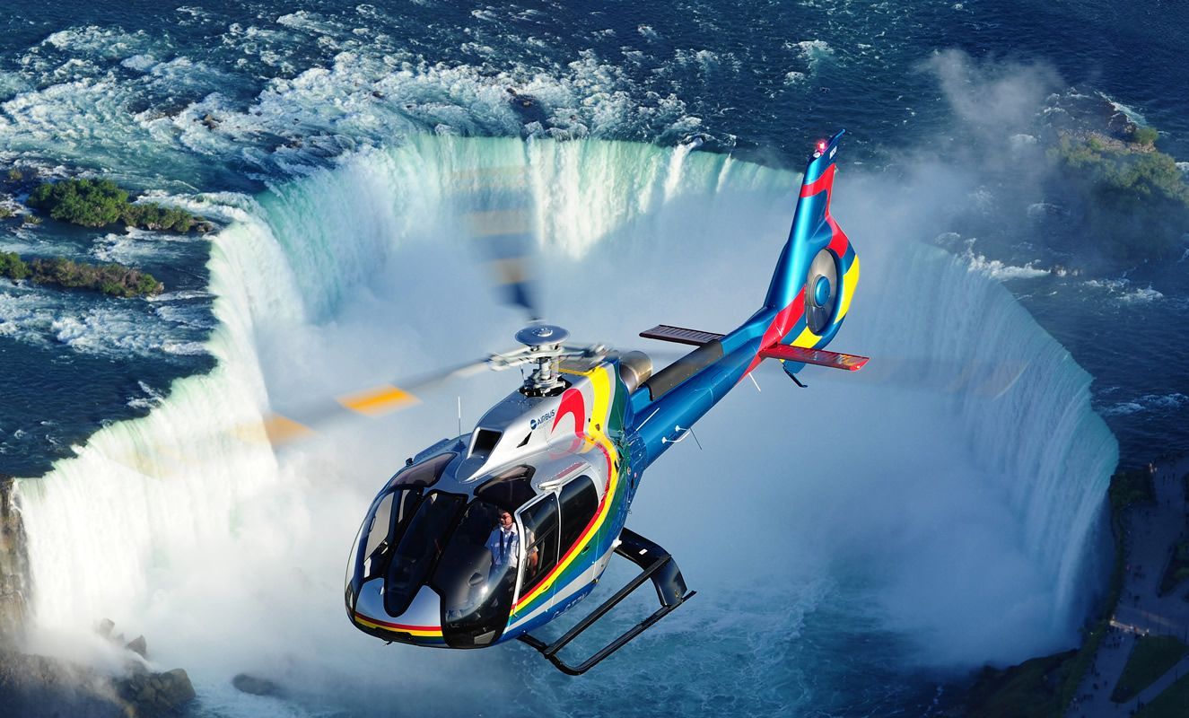 Een kleurrijke helikopter vliegt over een gigantische ijzige krater of gletsjer met kolkend water eronder.