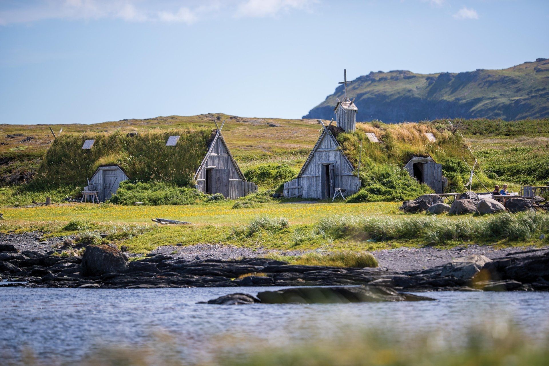 Kleine grijze Viking huisjes aan een grasrijke kustlijn met bergen en een blauwe lucht op de achtergrond.