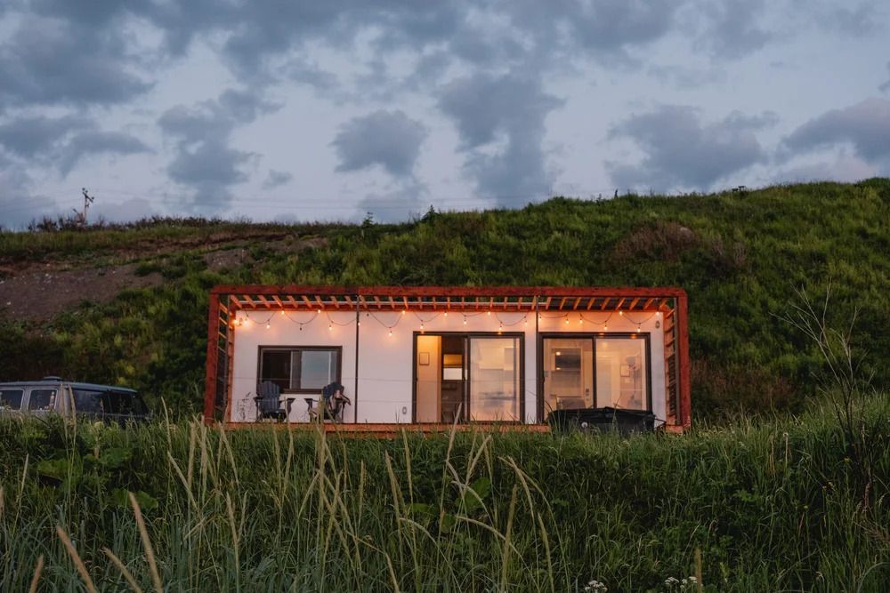 Moderne blokhut met warme verlichting, verscholen in het hoge gras op een heuvel bij schemering.
